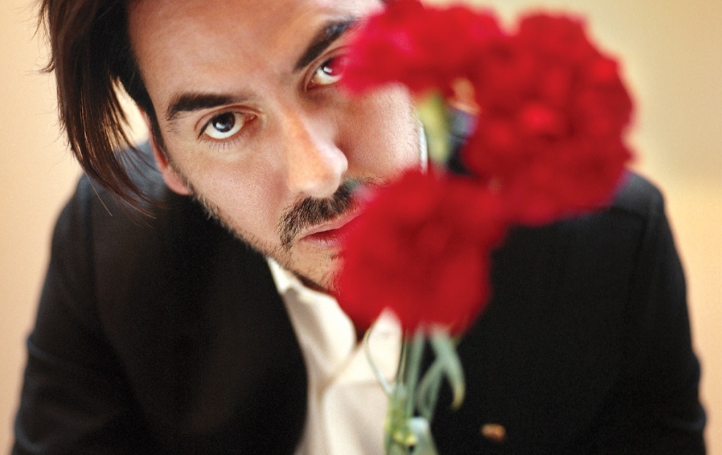 Image of Dhani Harrison &rsquo;01 in a black suit and white shirt, face partially obscured behind bright red flowers.
