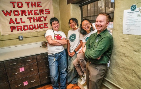 An image of Jo Ouyang, Yasmine Abdelaziz, Anna Ryu, and Michael Ziegler, with a sign in the background that states "to the workers all they produce."