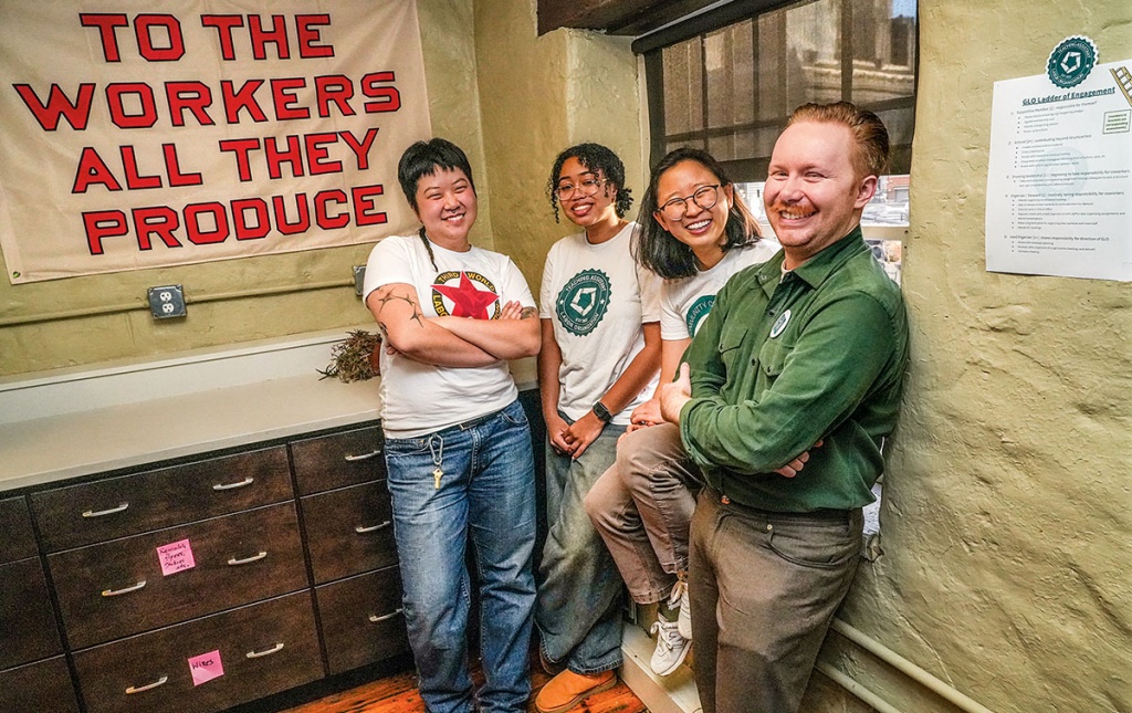 An image of Jo Ouyang, Yasmine Abdelaziz, Anna Ryu, and Michael Ziegler, with a sign in the background that states "to the workers all they produce."